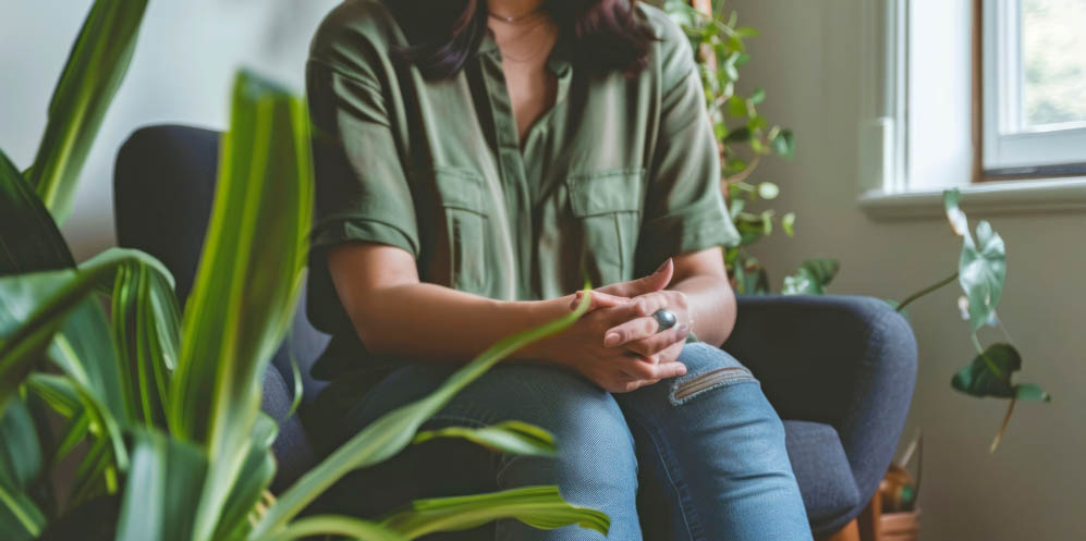 Image of a person wearing a casual green shirt and blue jeans. They are sitting in a chair by a window at the Distress Support Service with their hands clasped together.