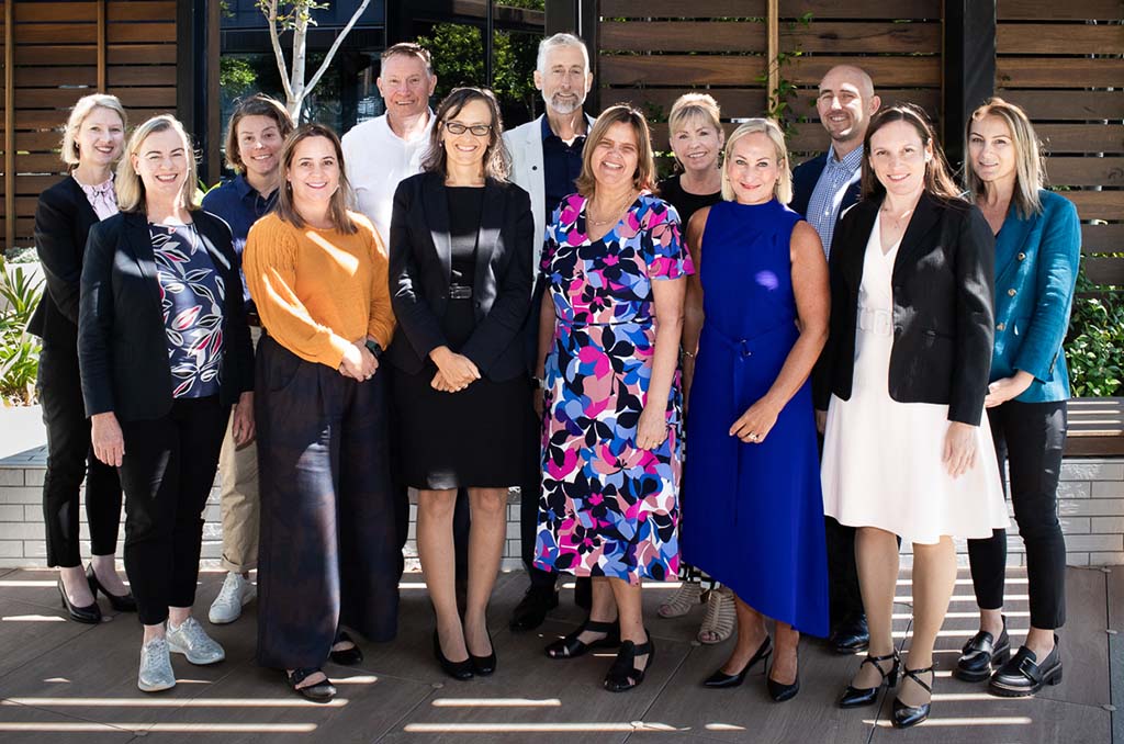 A group photo of 13 people standing outside in a sunny courtyard.