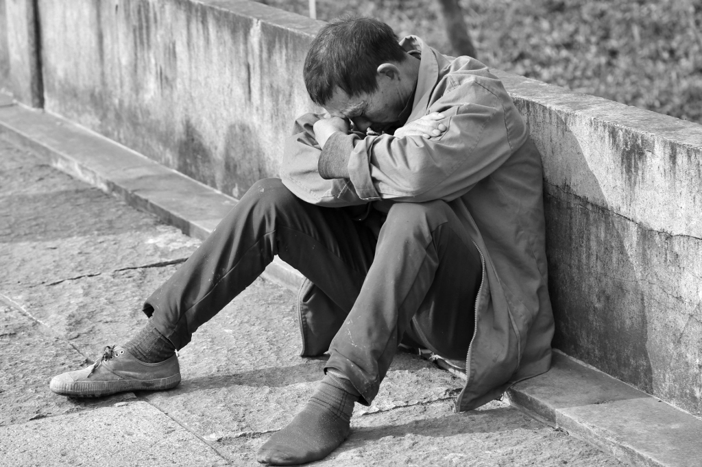 Image of a man sitting on the footpath resting his head on his arms.